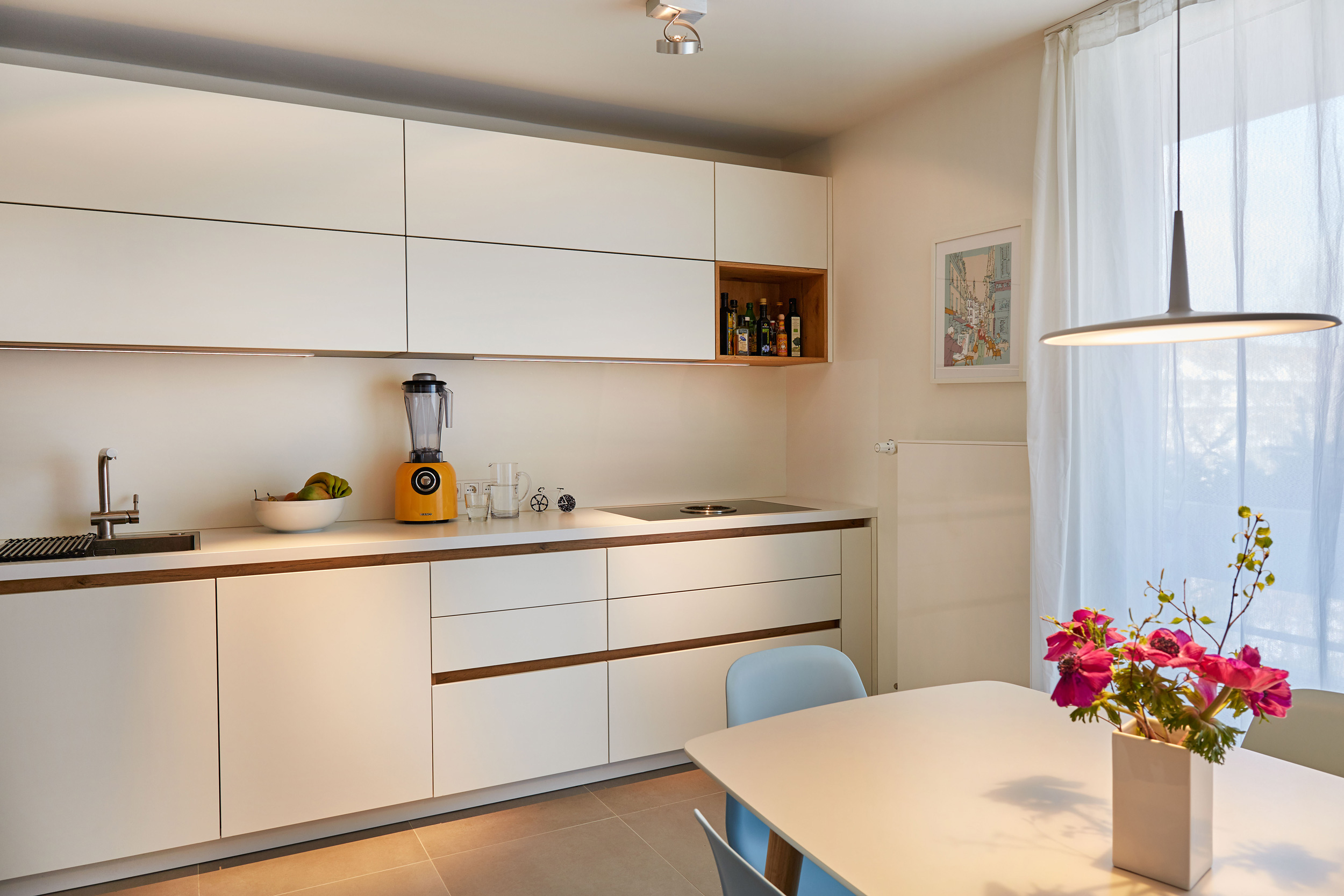 Foto: Modern gestaltete Wohnküche mit minimalistischer weißer Küchenzeile mit Holz Details, heller Esstisch mit hellblauen Stühlen und Pendelleuchte.