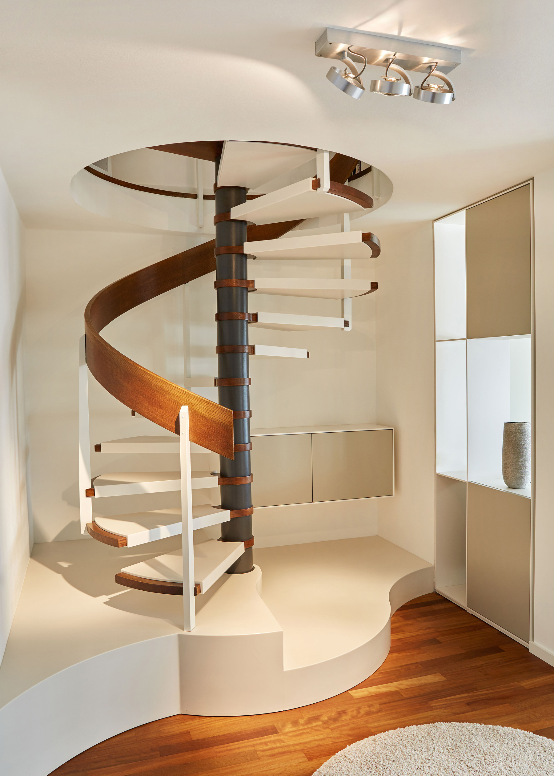 Foto: Moderner, heller Wohnraum mit weißer, skulpturaler Wendeltreppe mit Holzakzenten, warmem Parkettboden und minimalistischen Einbauschränken.