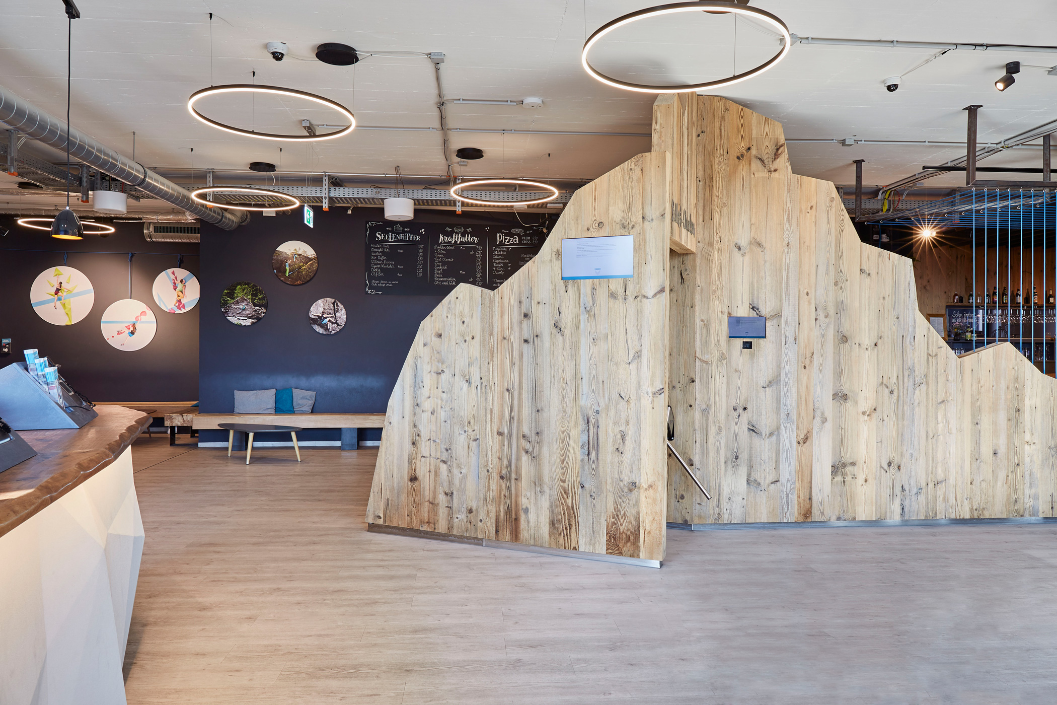 Foto: Modern gestalteter Empfangsbereich mit rustikalen Holzoberflächen, skulpturalem weißem Empfangstresen, raumtrennender Wand aus Altholz in Bergform und ringförmigen Lichtobjekten.