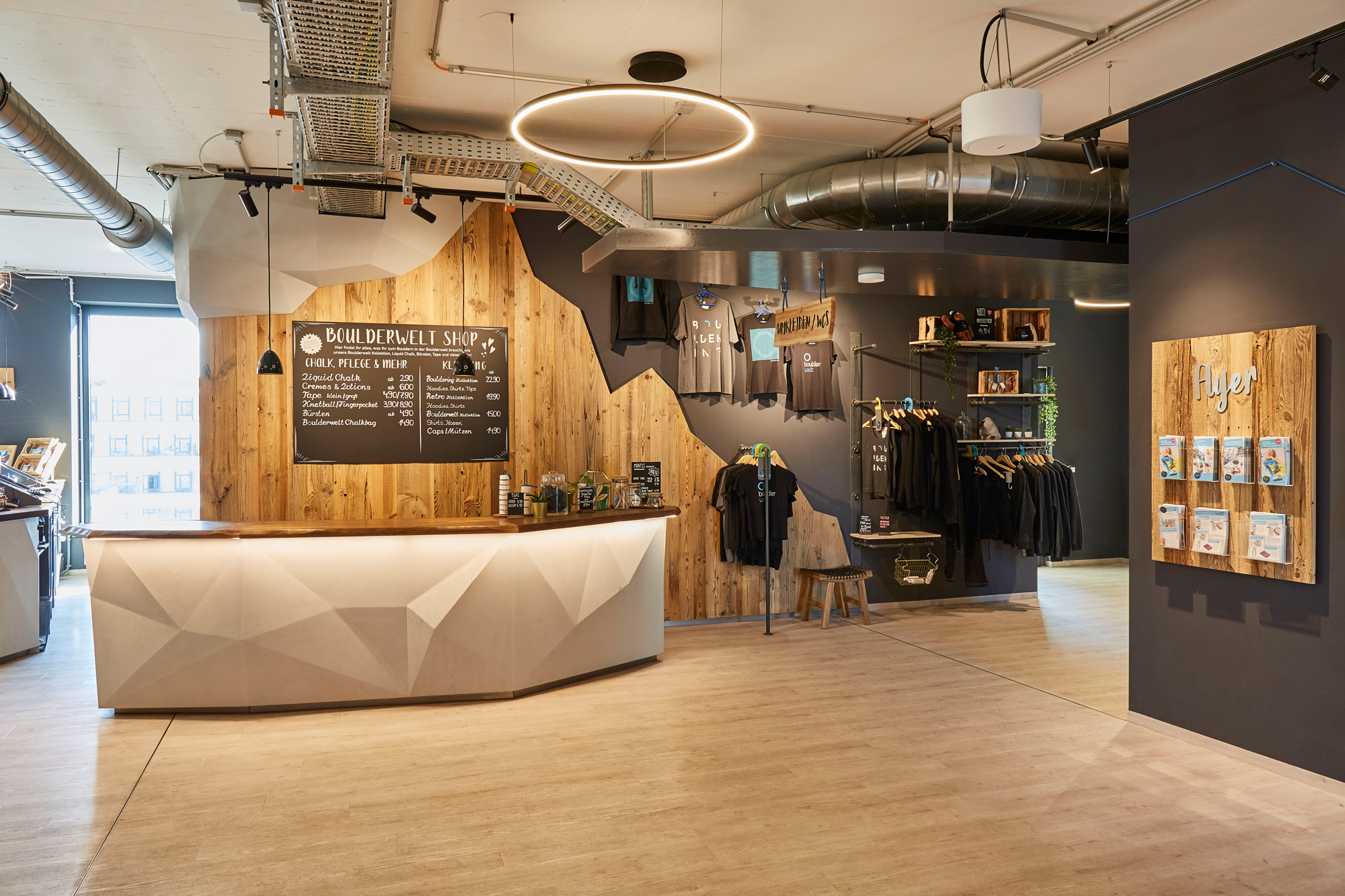 Foto: Empfangs- und Shopbereich mit geometrisch gestaltetem, weißen Tresen, Altholzoberflächen, integrierten Produktpräsentationen und moderner Beleuchtung in industrieller Architektur.