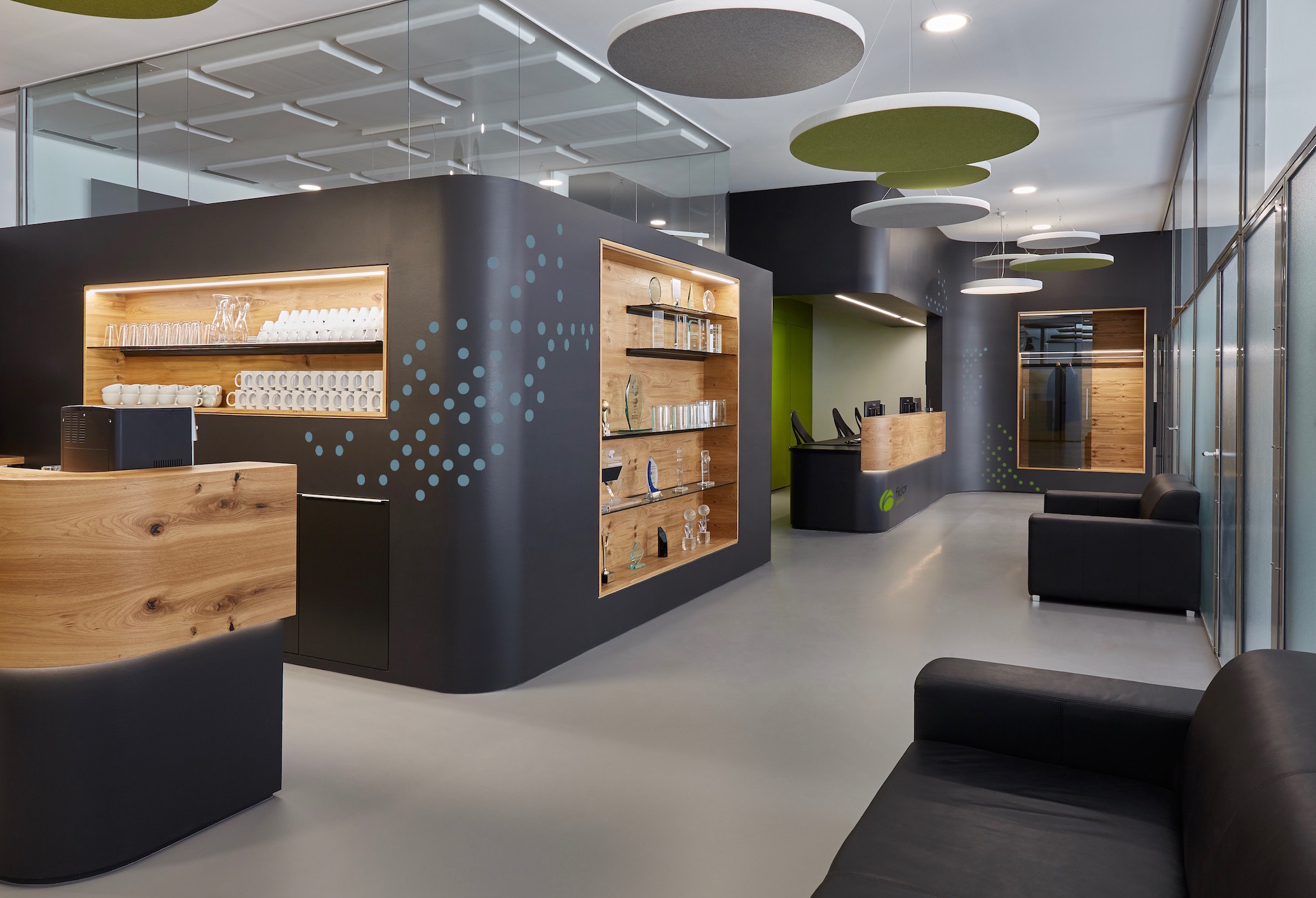 Foto: Empfangsbereich mit geschwungenen, schwarzen und holzverkleideten Einbauten, integrierten Regalen und Folierung in Punktmuster. Schwarze Sofas, grüne Akzente in Form von Wänden und runden Akustikelementen an der Decke. 