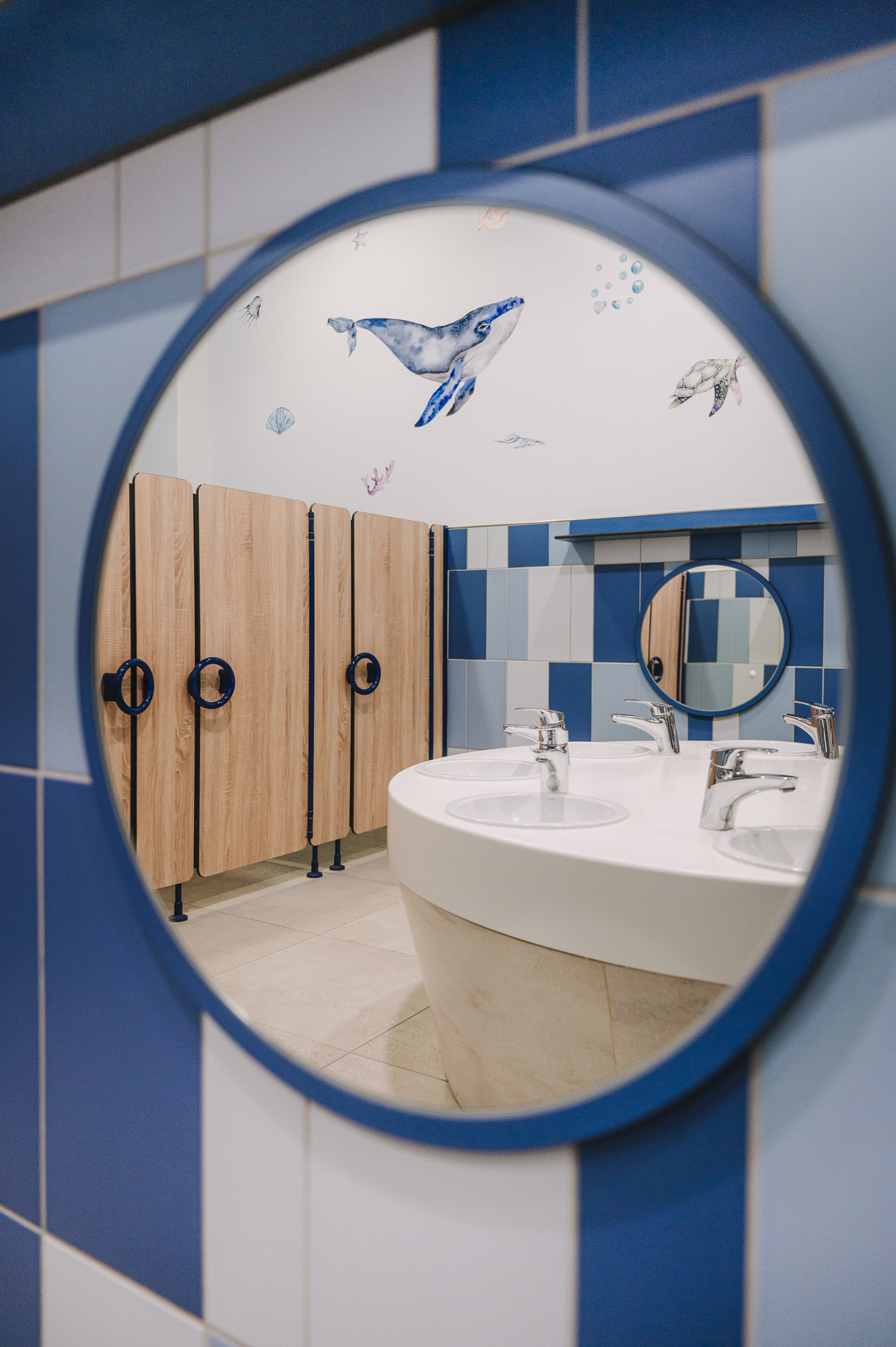 Foto: Detailaufnahme Spiegel, Spiegelung eines kinderfreundlich gestalteten Waschraums mit rundem Waschtisch, Holztrennwänden und maritimen Wandmotiven, blau-weiße Wandfliesen.