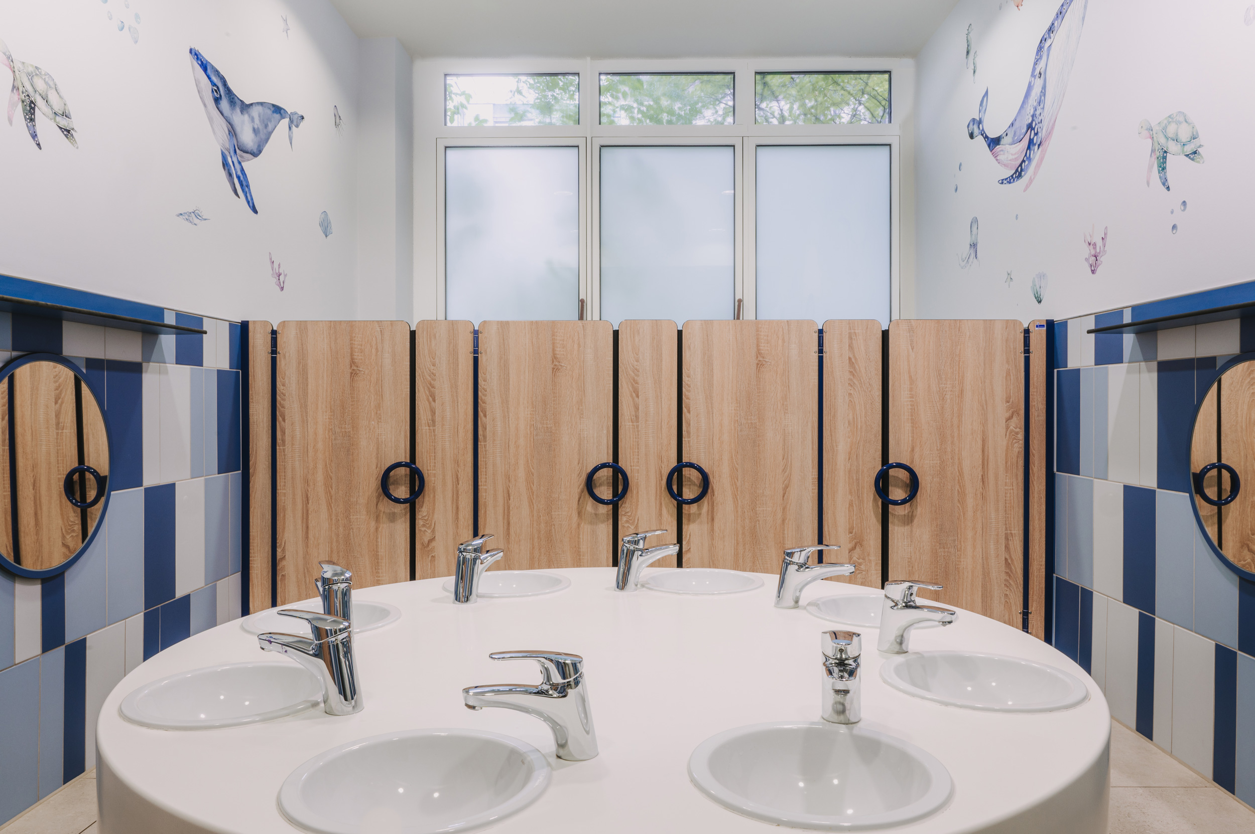 Foto: Symmetrisch gestalteter Waschraum mit ovalem Gemeinschaftswaschtisch, niedrigen Waschbecken, Holztrennwänden und blau-weißen Fliesen sowie maritimen Wandmotiven.