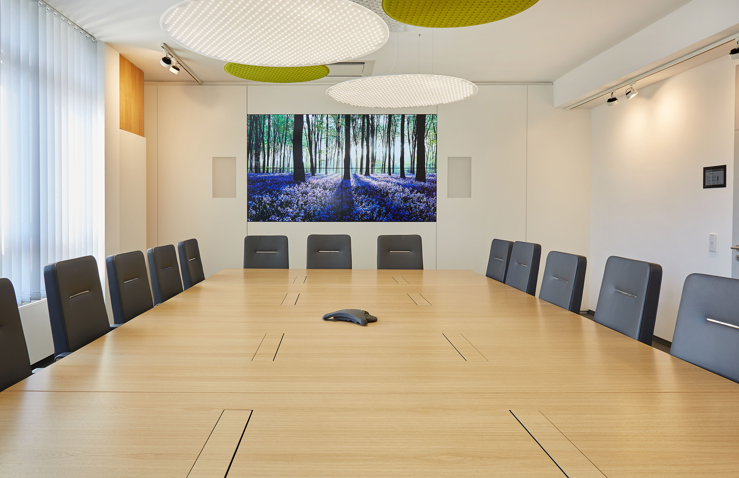 Foto: Moderner, heller Konferenzraum mit großem Holztisch, schwarzen Besprechungsstühlen, großem Bildschirm und runden grün-weißen Akustikelementen an der Decke.