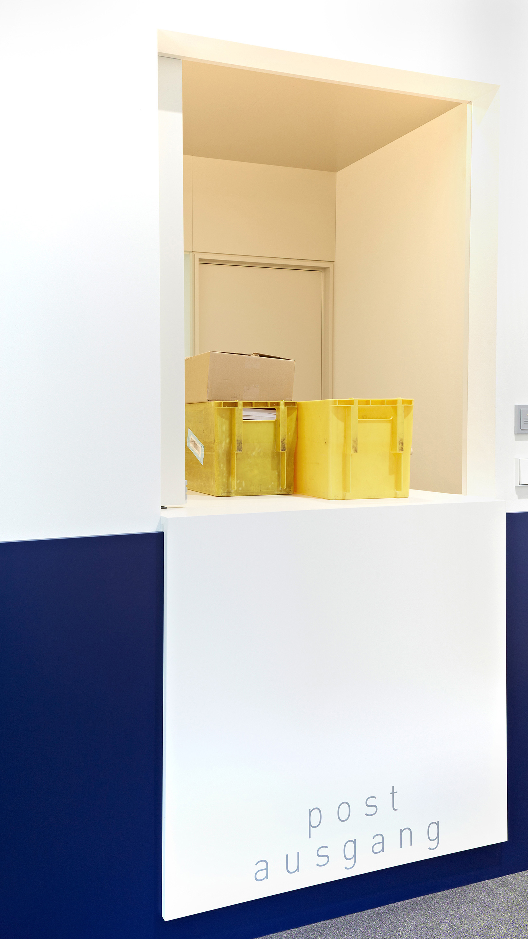 Foto: Detailaufnahme Poststelle Ausgangs Fenster mit gelben Postkisten und dunkelblauer Halbwand.