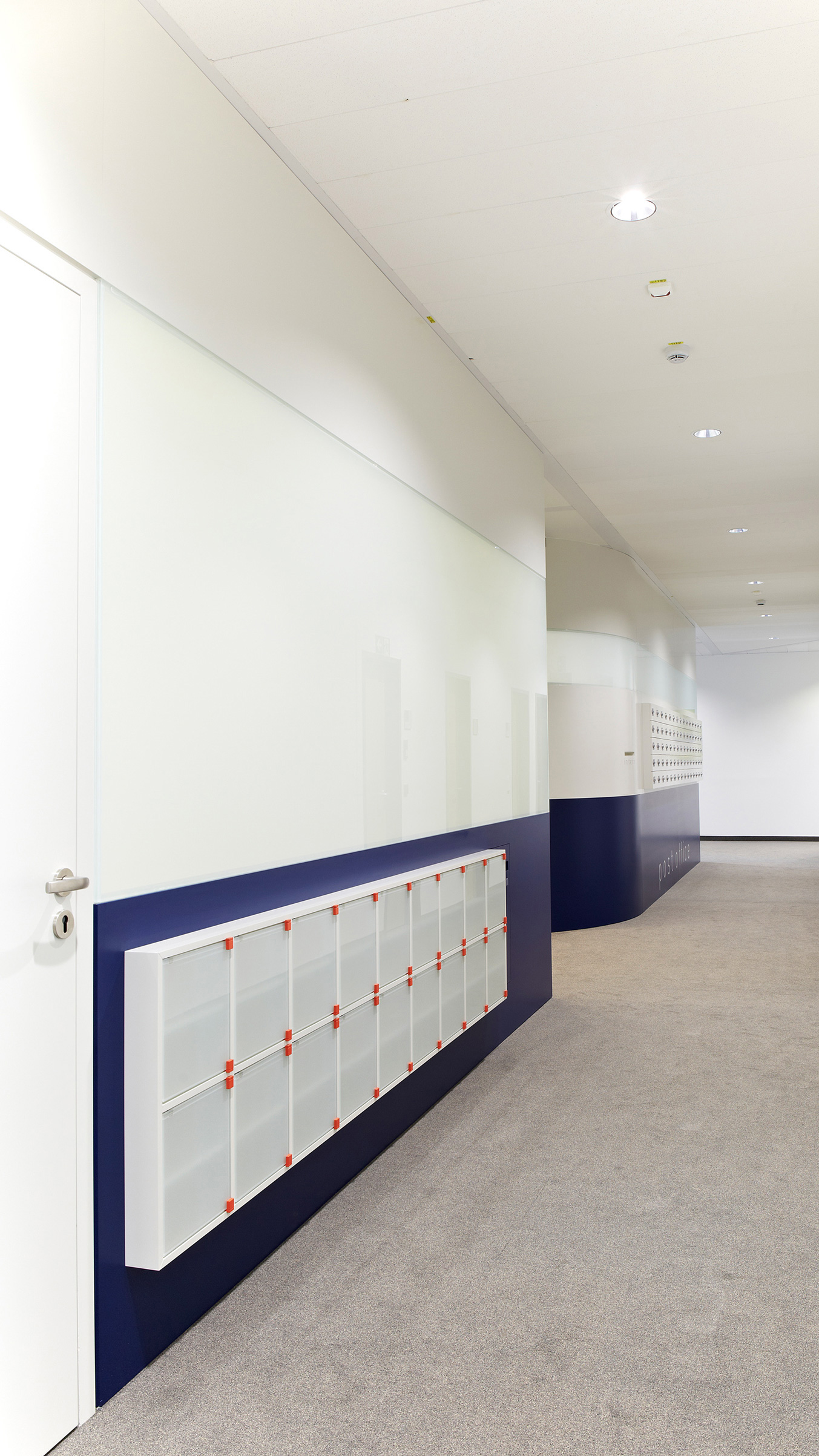 Foto: Flur einer modernen Post- und Paketstation in einem Bürogebäude mit weißen Fächern an der Wand, welche zur Hälfte dunkelblau gestrichen ist.