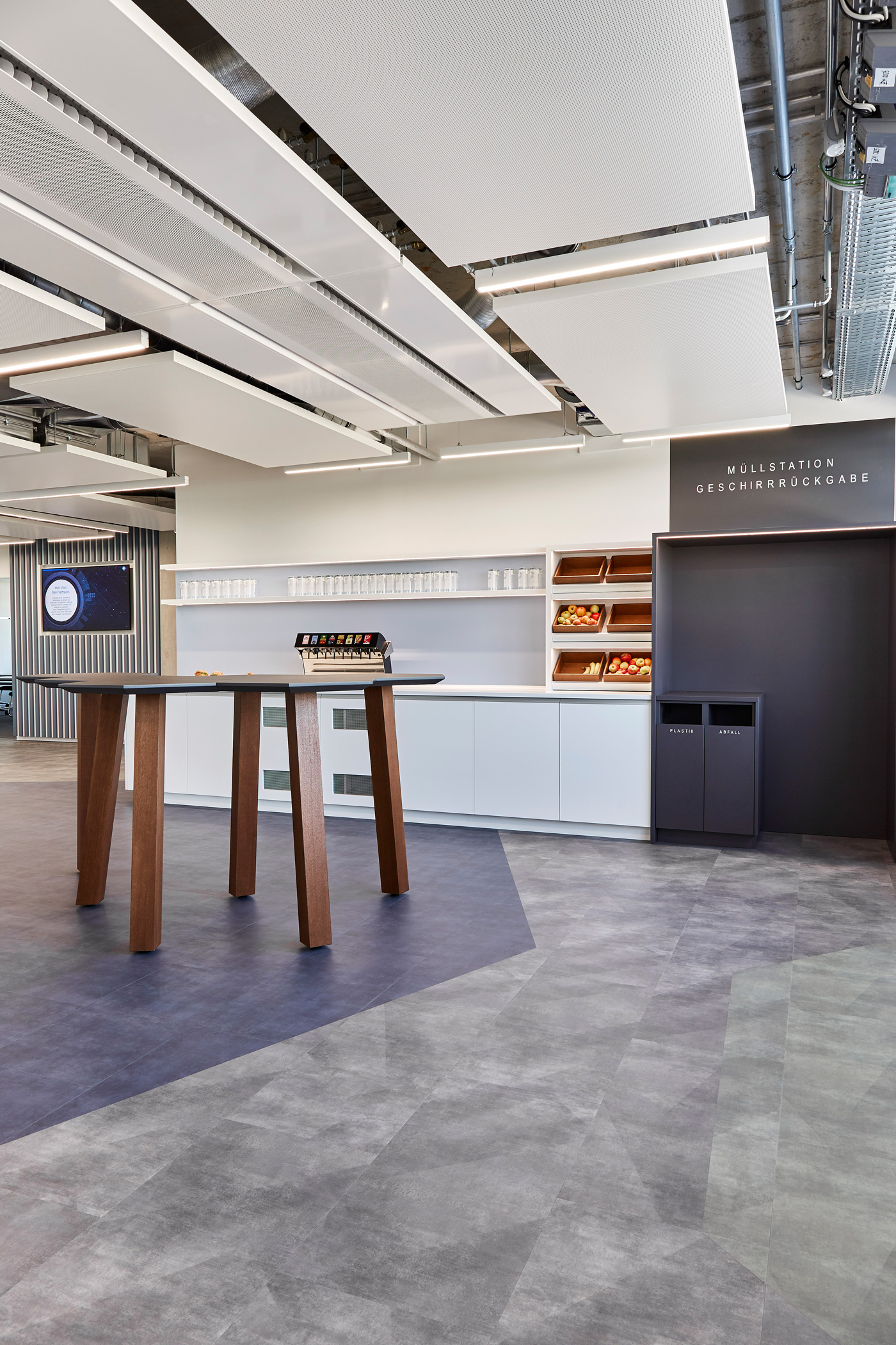 Foto: Moderne Teeküche mit weißer Küchenzeile, Obstauslage, Holz Stehtisch, dunkelgrauer Geschirrrückgabe und abgehängten Akustikpaneelen.
