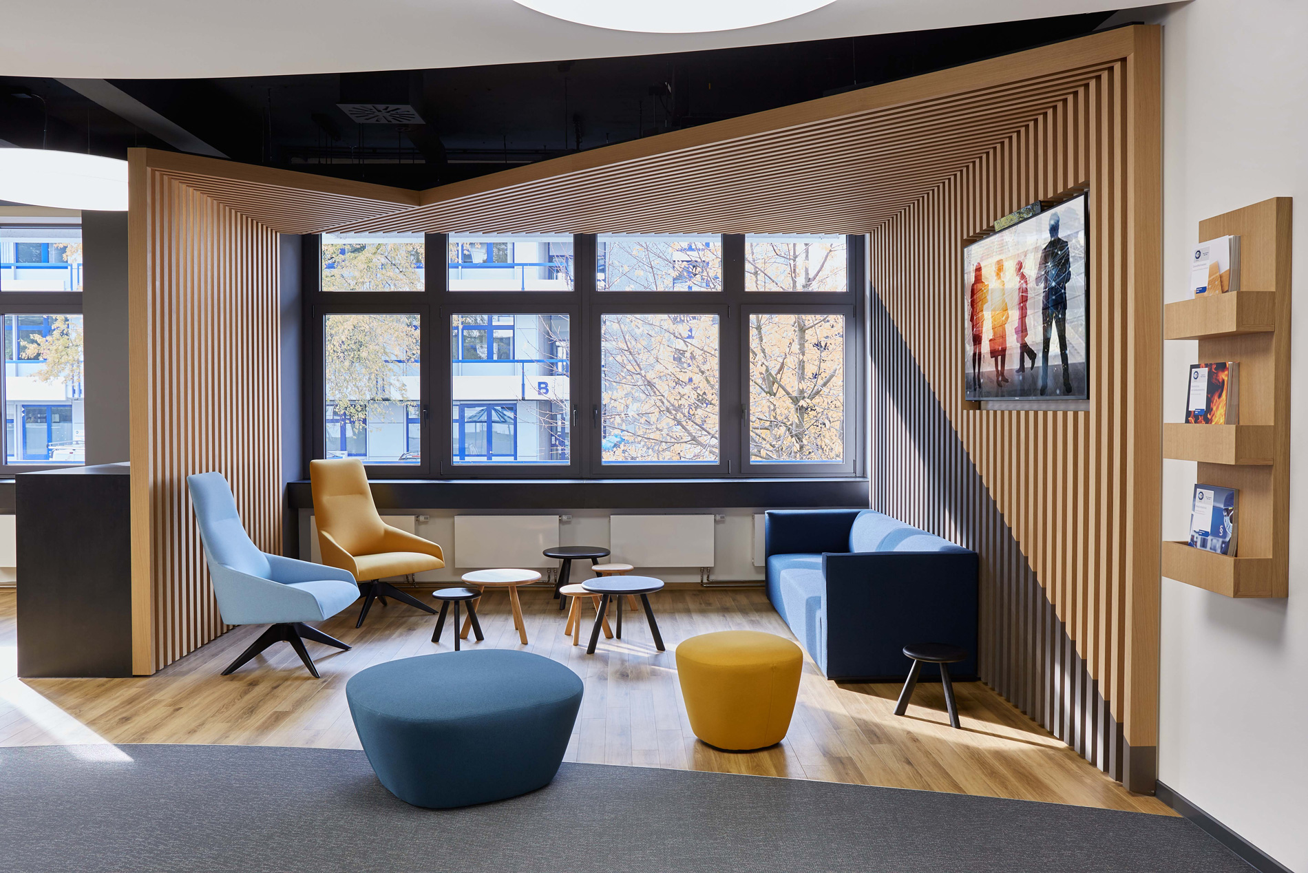 Foto: Moderne Lounge- und Kommunikationszone mit Sitzgruppe, gelb, blauen Sesseln und Hockern sowie einer markanten Holzlamellenkonstruktion und großen Fenstern.