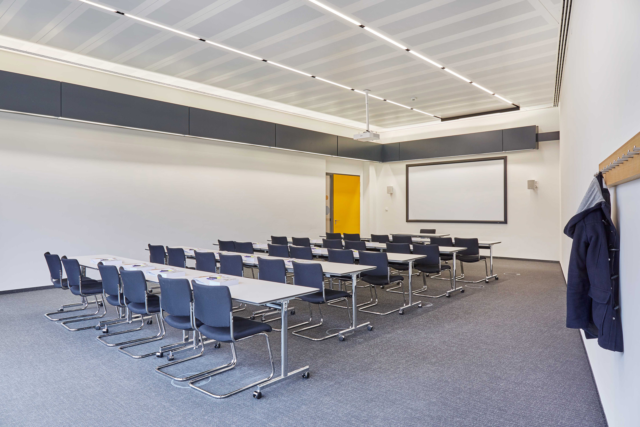 Foto: Moderner, heller Konferenz- oder Vortragsraum mit grauem Teppich, Garderobe, Leinwand und mehreren Tischreihen mit blauen Stühlen.