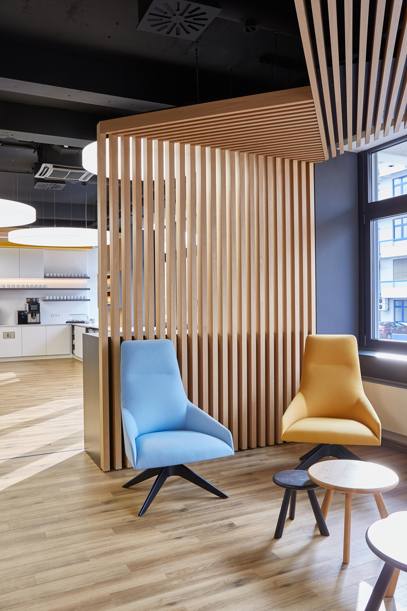 Foto: Moderne Lounge- und Kommunikationszone mit gelb, blauen Sesseln und Beistelltischen sowie einer markanten Holzlamellenkonstruktion und großen Fenstern.