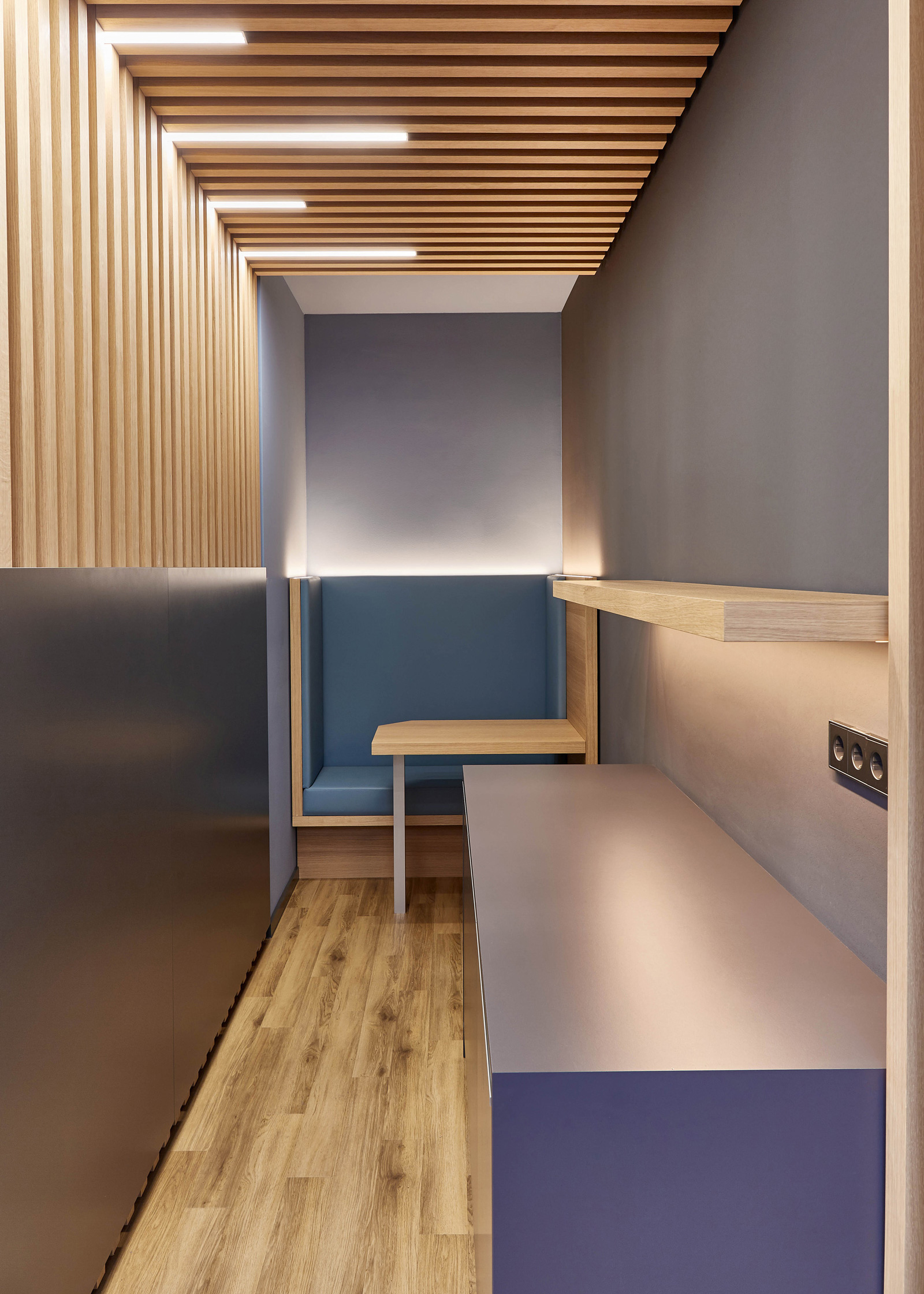 Foto: Moderner Innenraum mit Holzlamellen, integrierter Beleuchtung, blauer Sitznische, lila Arbeitsfläche und Holzfußboden in minimalistischer Gestaltung.