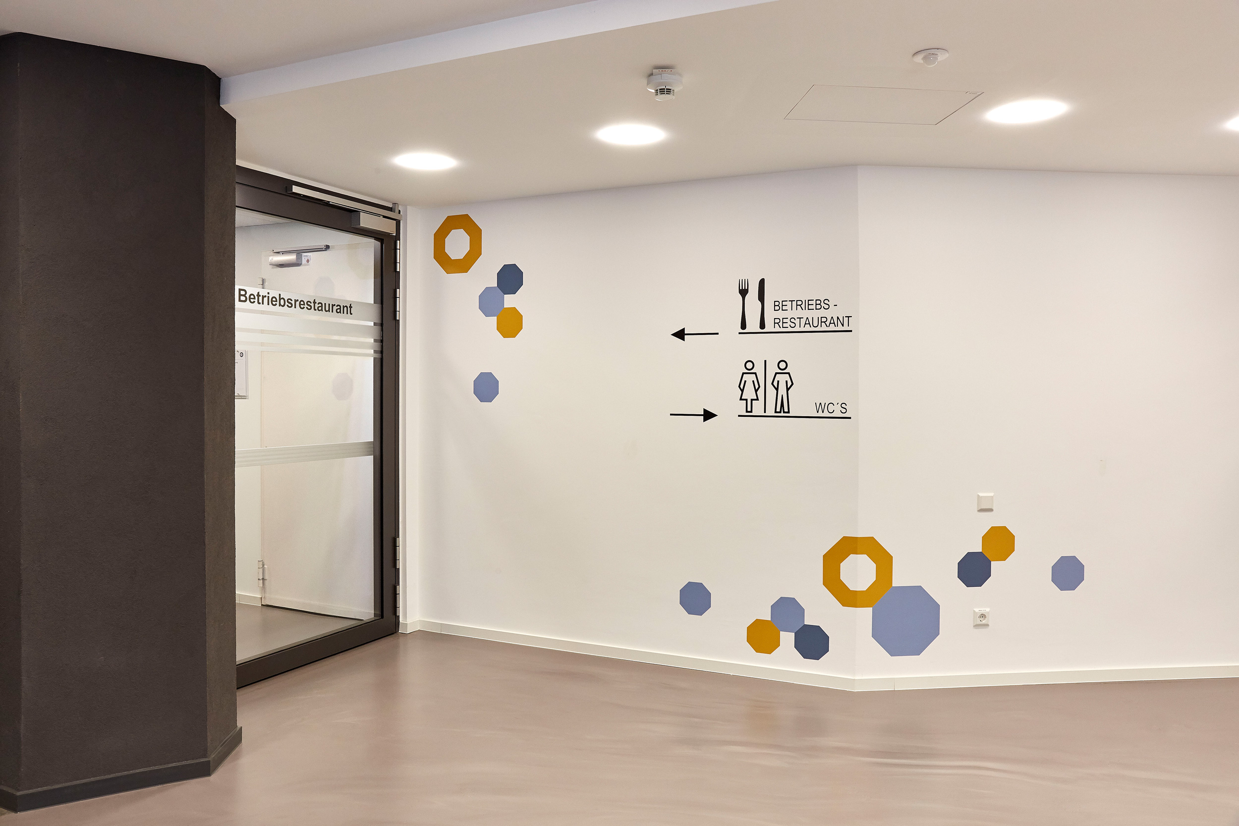 Foto: Modern, minimalistisch gestalteter Büroflur mit weißen Wänden, schwarzer Signaletik, sowie geometrische Folgerung in Gelb und Blau an den Wänden.