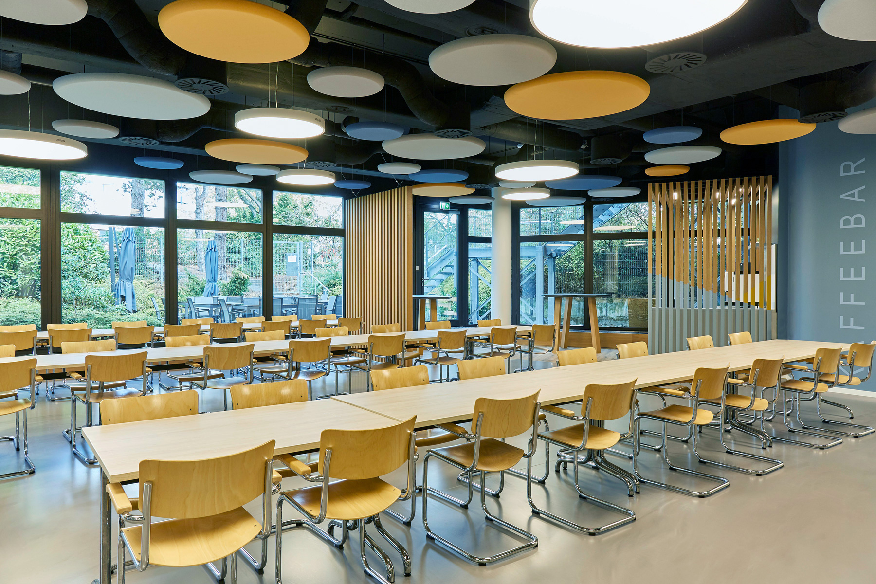 Foto: Modern gestalteter Kantinen- und Aufenthaltsbereich mit langen hellen Holztischen, zahlreichen Stühlen, großen Fensterfronten und runden Akustikelementen an der Decke in Weiß, Gelb und Blau. Helle Materialien, klare Linien und eine offene Raumgestaltung.