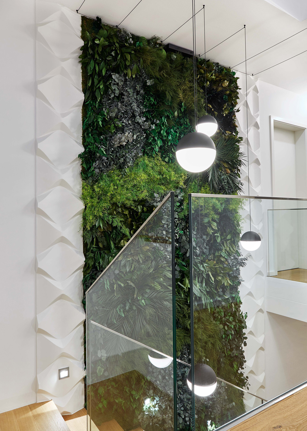 Foto: Moderne, offene Treppenhalle mit raumhoher Pflanzenwand, weißen 3D-Wandpaneelen, Glasgeländer und minimalistischen Pendelleuchten in Schwarz-Weiß.