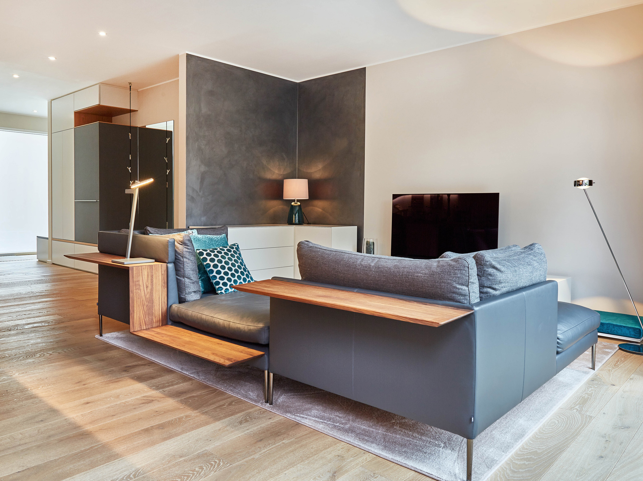 Foto: Modernes, offen gestaltetes Wohnzimmer mit grauem Designersofa, integrierten Holzablagen, hellem Holzboden, minimalistischer Beleuchtung und dunkelgrauer Akzent Wand in einer Ecke.