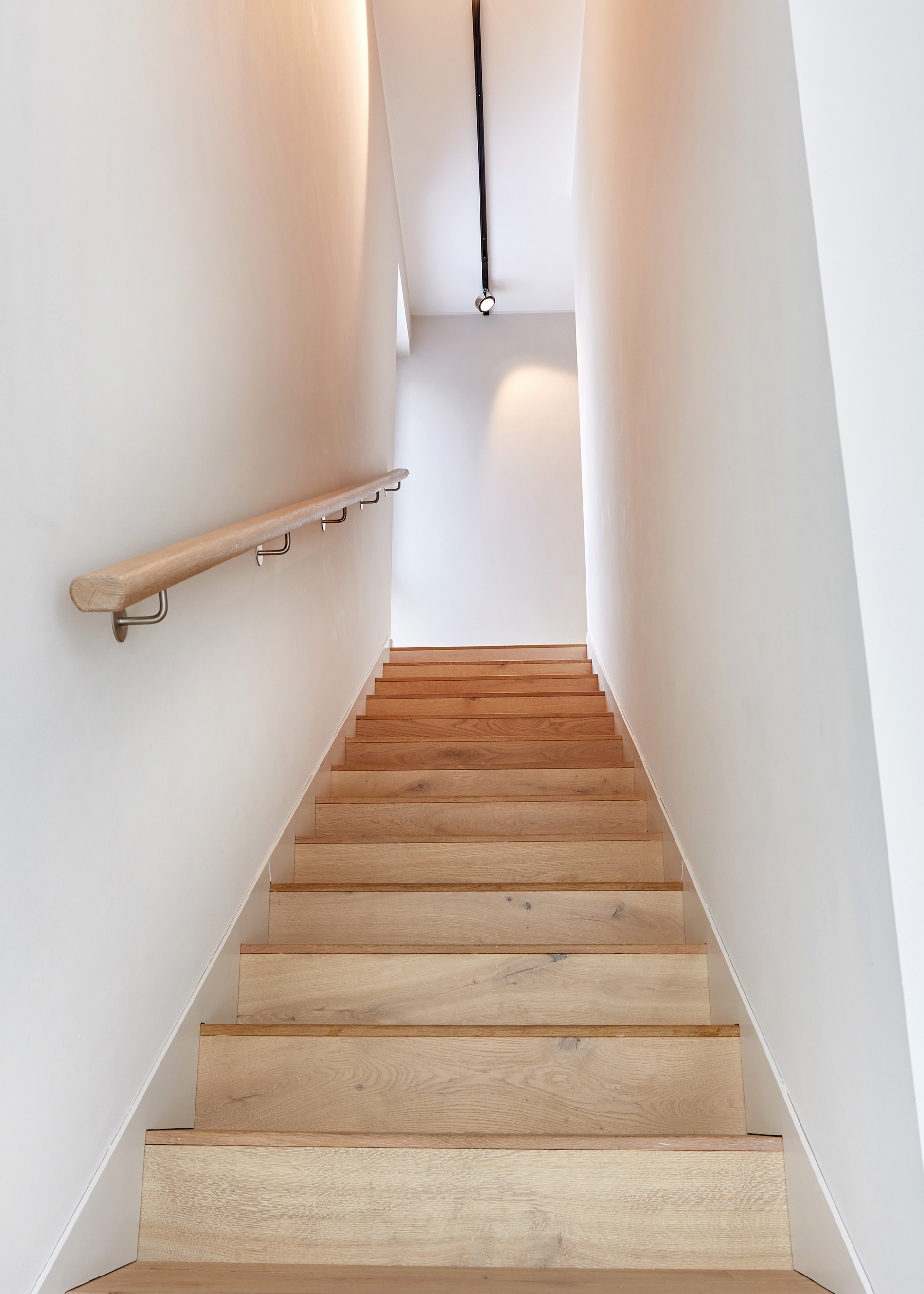 Foto: Schmale, gerade, minimalistische Treppe mit hellen Holzstufen, rundem Holz Handlauf und weißen verputzen Wänden.