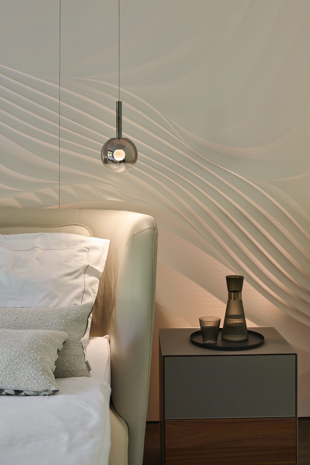 Foto: Detailaufnahme, Schlafzimmer Bettkopfteil aus hellem Leder, modernes Nachtkästechen in dunklem Holz und anthrazit, runde Glas-Pendelleuchten und weißen 3D-Wandpaneelen mit Wellen Optik.