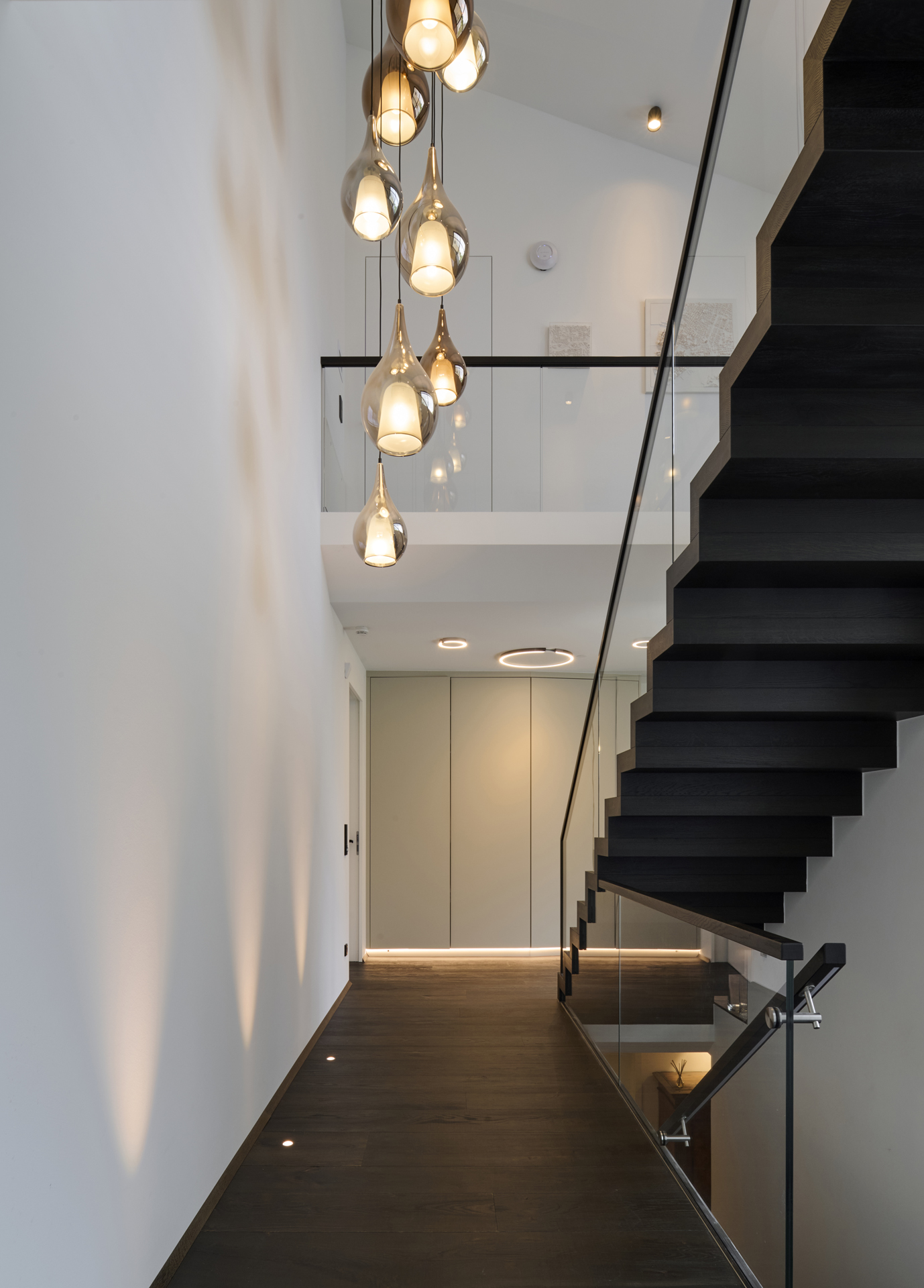 Foto: Moderner, lichtdurchfluteter Treppenaufgang  mit freitragender Holztreppe, Glasgeländer, hohen Decken, dunklem Holzboden und tropfenförmigen Akzent Pendelleuchten.