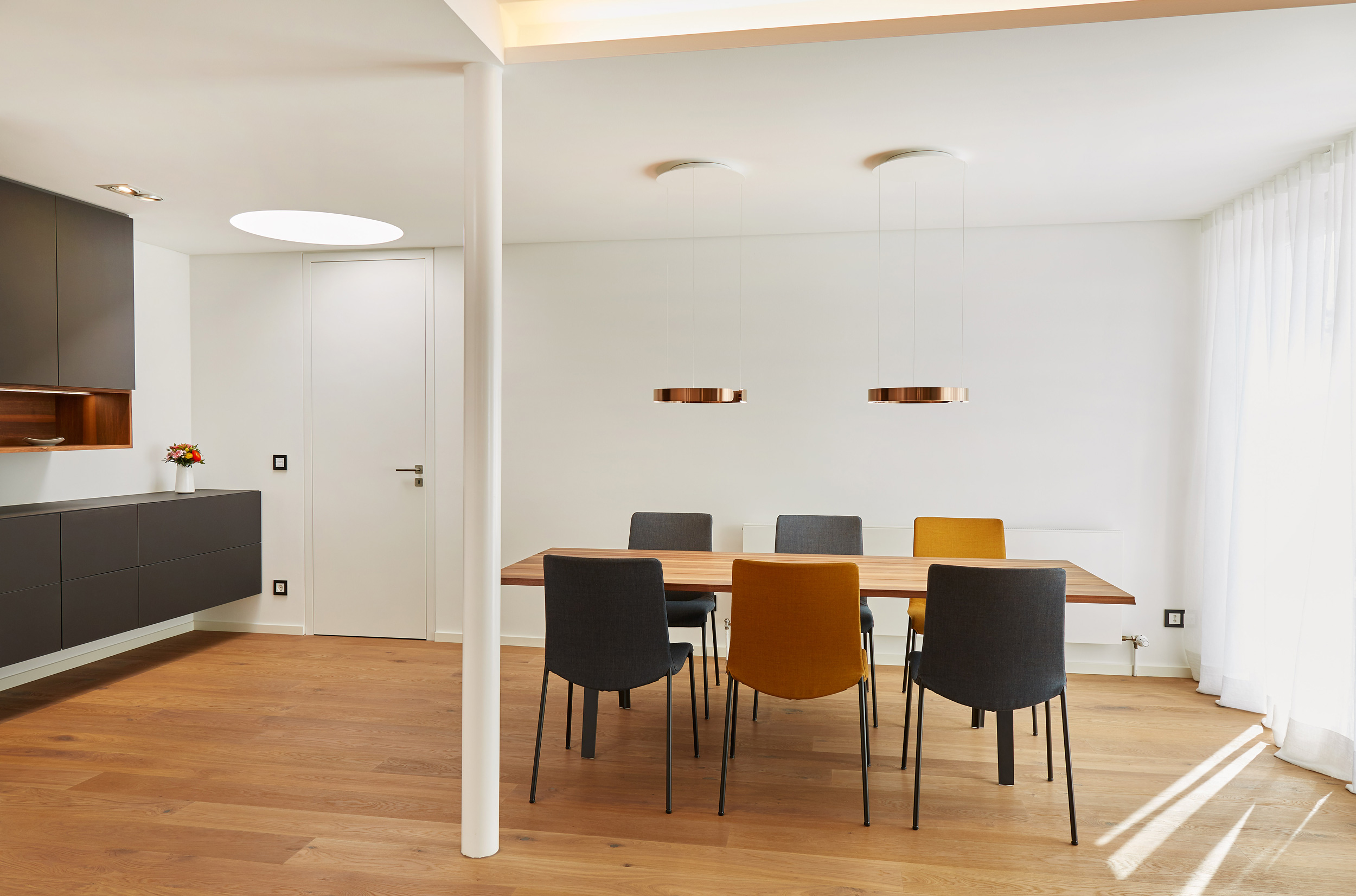 Foto: Offener, moderner, heller Wohn-/ Essbereich mit raumbildender Stütze, Holzesstisch, zweifarbigen Stühlen, modernen Pedelleuchten und dunklem, freihängendem Sideboard.