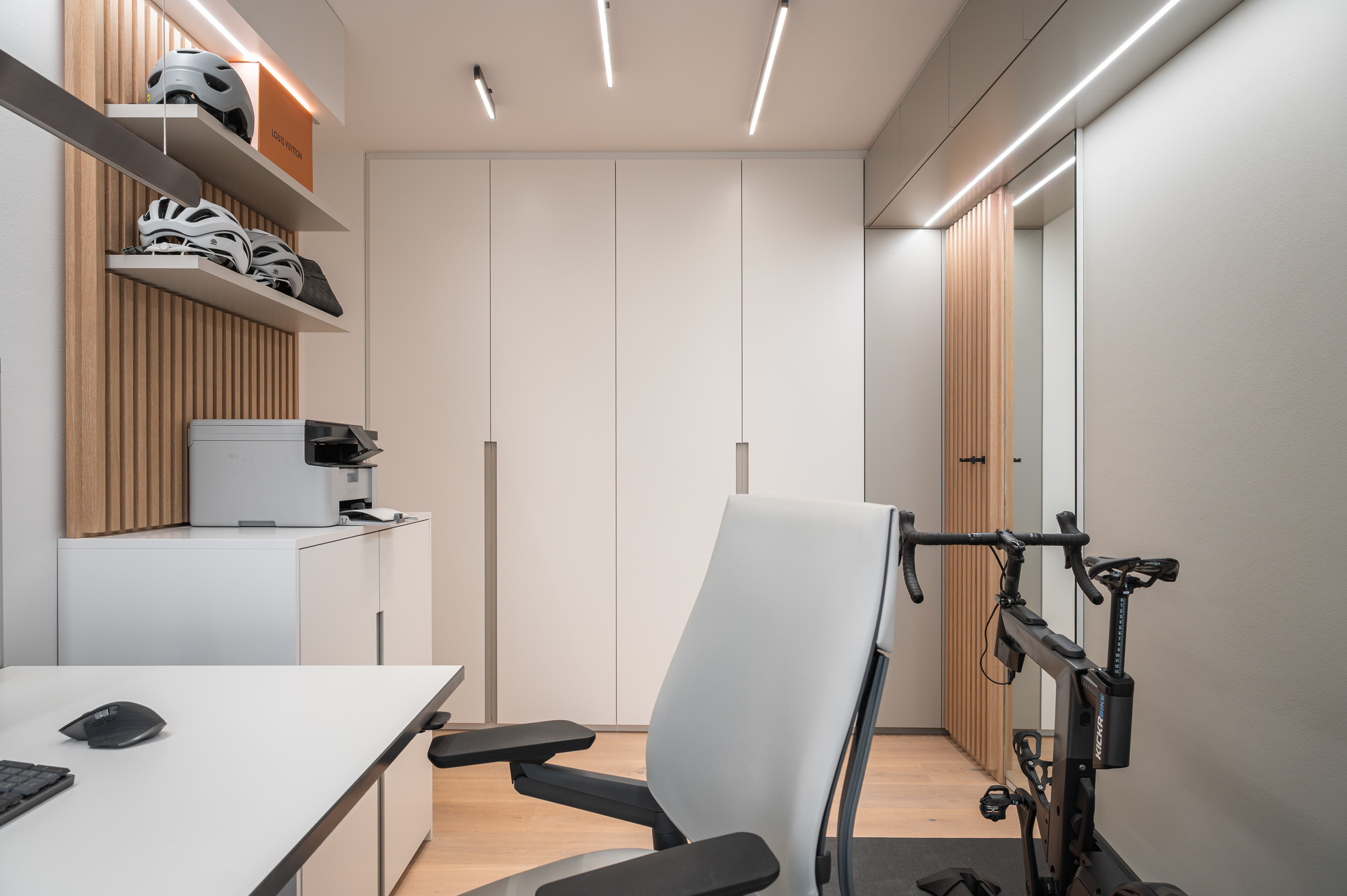 Foto: Kompaktes, modernes Homeoffice mit weißen Einbaumöbeln, Holzlamellen, linearer Deckenbeleuchtung und Indoor-Fahrrad neben dem Schreibtisch.