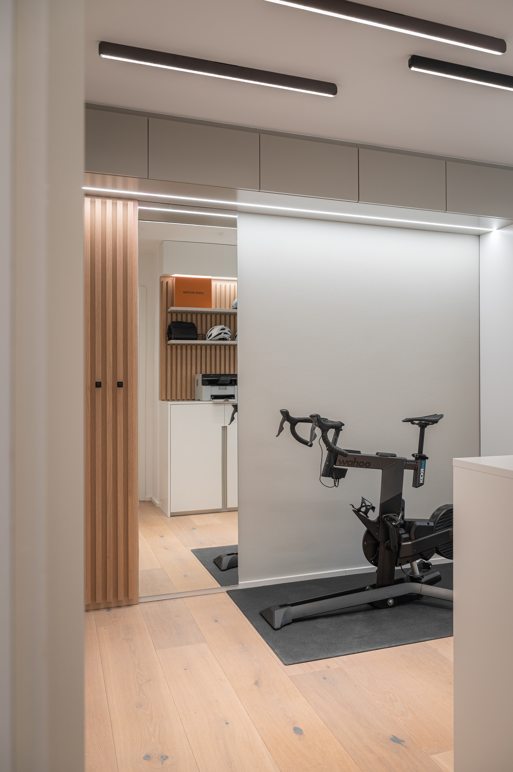 Foto: Kompaktes, modernes Homeoffice mit weißen Einbaumöbeln, Holzlamellen, integriertem Spiegel, linearer Deckenbeleuchtung und Indoor-Fahrrad.